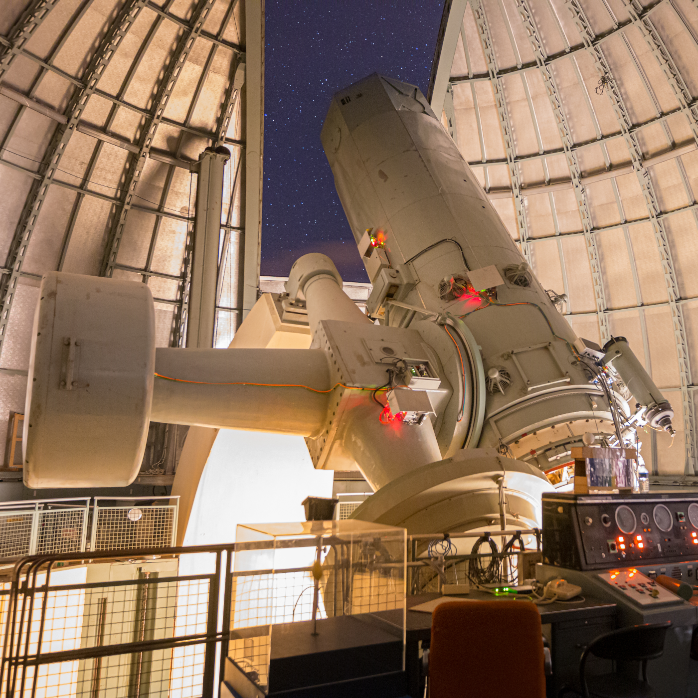 Un télescope sous une coupole ouverte. On voit les étoiles dehors. À l'avant-plan, il y a une console.