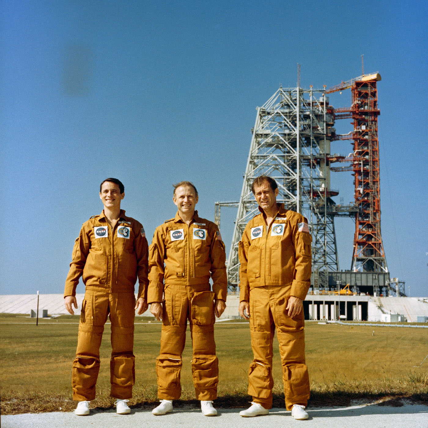 Trois hommes en combi orange devant un pas de tir de fusée.